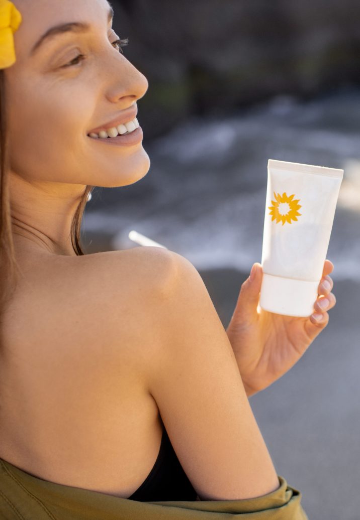 Beautiful young woman with hairflower holding tube with sunscreen lotion while resting on the beach on a summer vacations. Sun protection concept for skin